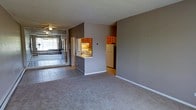 This single-floor residence offers a practical and functional layout centered around an open-concept living space. The main living area is finished with wall-to-wall carpeting and neutral gray walls, creating a calm and understated atmosphere throughout. A ceiling fan overhead adds both comfort and a subtle design touch to the room.

The kitchen is efficiently designed with warm wood-toned cabinetry and features a pass-through counter that connects it to the living area, promoting an open and communicative flow between spaces. White appliances, including a refrigerator and a range, are neatly integrated into the layout, and under-cabinet lighting adds a functional brightness to the workspace.

Adjacent to the main living area is an enclosed sunroom or patio space, separated by large mirrored sliding glass doors. This bright extension is finished with tile flooring and benefits from generous natural light streaming through its windows, offering a versatile space that can serve as a lounge, reading nook, or additional entertaining area.

The transition between the tiled entryway and the carpeted living room adds a natural visual delineation between zones. Overall, the space presents a straightforward and livable layout with clean lines and a neutral palette well-suited for a variety of furnishing styles.