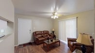 This single-floor space presents a functional and comfortable living area with several notable features. The living room is anchored by warm hardwood-style flooring that extends throughout, adding a sense of cohesion and warmth to the space. A ceiling fan with a built-in light fixture is centrally mounted, providing both air circulation and ambient lighting to the room.

Natural light is welcomed through a large sliding glass door dressed with white vertical blinds, offering the ability to control light levels while maintaining privacy. The entryway features a solid white door with standard hardware, conveniently located adjacent to the living area.

The walls are finished in a neutral off-white tone, complementing the textured ceiling and creating a versatile backdrop suitable for a variety of décor styles. The living area comfortably accommodates a reclining loveseat and matching armchair alongside a dark marble-topped coffee table, demonstrating the practical use of the available space. A floor-to-ceiling baseboard heating unit is also visible, indicating a dedicated climate control system. Overall, the space presents a straightforward, functional layout with practical finishes throughout.