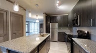 This contemporary single-floor residence showcases a thoughtfully designed open-concept layout that seamlessly connects the kitchen and living areas. The kitchen is a standout feature, appointed with rich dark wood cabinetry that provides striking contrast against light granite countertops. A generous island with an undermount sink serves as both a functional workspace and a social hub, illuminated by elegant pendant lighting overhead. Stainless steel appliances, including a full-size refrigerator and range, lend a polished, professional feel to the space. A tasteful tile backsplash adds texture and visual interest along the walls. Track lighting along the ceiling ensures the workspace is well-lit throughout. Light-toned flooring runs continuously through the kitchen and into the adjacent living area, enhancing the sense of openness and flow. The living space benefits from expansive floor-to-ceiling windows that invite generous amounts of natural light and offer views of the surrounding greenery, creating a bright and welcoming atmosphere. The overall design balances modern aesthetics with practical functionality, resulting in a refined and comfortable living environment.
