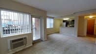 This single-floor unit offers an open and functional layout that seamlessly connects the living area to the kitchen. The living space is finished with wall-to-wall carpeting and painted in warm, neutral tones that contribute to a welcoming ambiance. Natural light flows generously through large windows fitted with vertical blinds, as well as a glass door that opens to an exterior balcony, offering views of the surrounding brick building exterior. A wall-mounted HVAC unit provides climate control directly within the living area.

The kitchen is positioned at the back of the open floor plan and is accessible via a raised breakfast bar counter, which serves as a natural dividing element between the two spaces. The kitchen features dark cabinetry, a stainless steel microwave and range, a white refrigerator, and overhead lighting that illuminates the workspace efficiently. The raised counter provides an opportunity for casual dining or additional workspace.

Overall, the unit presents a clean, straightforward layout well-suited for everyday living. The combination of natural light, open sightlines between the living and kitchen areas, and practical finishes makes this a functional and comfortable single-floor space.