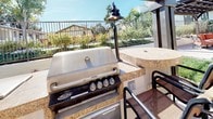 This single-level outdoor communal space is thoughtfully designed to encourage relaxation and social interaction in an open-air setting. The layout is anchored by multiple pergola structures with dark wood beam detailing, which provide partial shade over bar-height seating areas and an outdoor kitchen station. The outdoor kitchen is equipped with a stainless steel built-in Turbo gas grill set into granite-topped counters, accompanied by pendant-style lighting that adds a refined touch to the functional setup.

The patio extends generously across a smooth concrete surface, offering multiple seating zones furnished with wrought-iron tables and chairs beneath large orange market umbrellas. A raised, stone-clad planter bed filled with drought-tolerant succulents and ornamental plantings serves as a natural focal point near the main gathering area. Mature trees frame the perimeter, providing a green backdrop and filtered shade throughout the day.

The surrounding landscaping includes manicured lawn areas and potted palms that add texture and warmth to the hardscape. The overall ambiance is bright, airy, and well-maintained, creating an inviting environment well suited for outdoor dining, casual gatherings, and everyday leisure.