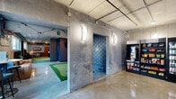 Located at 127 N 17th St in Richmond, Virginia, this single-floor commercial entertainment space offers a unique indoor golf experience with a distinctly industrial aesthetic. The venue is defined by its raw concrete walls, polished concrete floors, and exposed ceiling infrastructure, creating an urban, warehouse-inspired atmosphere throughout.

The main area opens to a well-appointed lounge zone featuring a tan leather sofa, bar-height seating, and street-facing windows that flood the space with natural light and views of the surrounding neighborhood. A branded "GOLF" wall installation adds to the playful, golf-centric character of the venue. An indoor putting green with multiple holes and flag markers runs through the central area, offering casual play for guests.

A dedicated golf simulator bay provides an immersive experience, complete with a large projection screen, turf mat hitting area, and acoustic foam paneling on surrounding walls for sound management. Blackout curtains enclose the simulator area for optimal visibility during play.

Near the entrance, a retail and checkout station features snack shelves and refrigerated beverage displays, providing convenient refreshments for guests. Decorative blue diamond-patterned wallpaper accents select walls, adding a pop of color and personality against the raw concrete backdrop. A welcoming bench seat near the entrance serves as a comfortable waiting area, completing this thoughtfully designed golf entertainment venue.