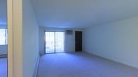 This single-floor space centers around a generously sized main living area finished with wall-to-wall carpeting that extends throughout the room. A large sliding glass door dominates one wall, fitted with vertical blinds that allow for adjustable natural light and provide access to an outdoor area. The bright white walls and light-toned ceiling contribute to an open, airy feel within the space. A wall-mounted air conditioning unit is positioned above the sliding door, offering a built-in climate control solution. A dark-toned entry door is situated adjacent to the sliding glass door, providing a private point of entry. The room's layout is open and unobstructed, offering flexibility for various furniture arrangements. Electrical outlets are distributed along the walls, ensuring practical functionality throughout. The overall ambiance is clean and straightforward, with neutral finishes that serve as a blank canvas for personalization.