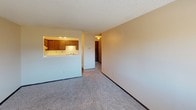 This single-floor residence offers a practical and straightforward layout, as revealed by the 3D floor plan model. The living area is generously proportioned, featuring neutral-toned carpet, beige walls, and a textured popcorn ceiling. A wall-mounted HVAC unit and baseboard heating run along the perimeter, ensuring climate control throughout. Windows fitted with horizontal blinds allow natural light to filter in, offering views of surrounding outdoor greenery.

The kitchen is connected to the living space via a pass-through counter opening, providing an open, communicative feel between the two areas. The kitchen itself is equipped with rich, dark wood cabinetry offering ample storage, white laminate countertops, a coil-burner electric range with a range hood above, and a dishwasher. Dark wood flooring grounds the kitchen space.

The bedroom is well-sized with the same carpeted flooring and textured ceiling found throughout. A closet with a dark wood door provides dedicated storage, and a window brings in natural light.

The bathroom features a full tub and shower combination, finished with clean white square tile surround and chrome fixtures, maintaining a simple and functional aesthetic consistent with the rest of the unit.