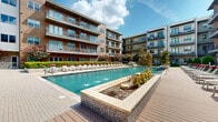 Located at 3900 W Northwest Hwy in Dallas, Texas, this multi-story residential community is defined by its striking contemporary architecture, blending warm wood-tone cladding, stone accents, and stucco exteriors across its two floors and beyond. The centerpiece of the property is a expansive resort-style swimming pool set within a beautifully landscaped courtyard, surrounded by rows of lounge chairs and raised planter beds featuring mosaic tile detailing and water features that add a dynamic, resort-like atmosphere.

The outdoor amenity area is thoughtfully designed for both relaxation and social gathering. A spacious pool deck extends into a dedicated entertaining terrace equipped with dining tables shaded by bold red umbrellas, outdoor grilling stations, and a fire feature surrounded by additional lounge seating. Tucked into a lush garden corner, a private lounge nook offers wicker daybeds with red cushions, framed by mature trees and colorful plantings for a more secluded retreat.

Residents benefit from private balconies and patios on multiple levels, offering direct views of the courtyard and greenery below. The well-manicured grounds incorporate a mix of flowering trees, sculpted shrubs, and open lawn areas, creating a vibrant and inviting outdoor environment that complements the community's modern architectural aesthetic.