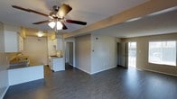 This single-story space features an open-concept layout that seamlessly connects the main living area to the kitchen, creating a functional and versatile floor plan. Dark hardwood-style flooring runs throughout, providing a clean contrast against the neutral, light-toned walls and white baseboards.

The living area is generously sized and anchored by a ceiling fan with an integrated light fixture, offering both comfort and illumination. A textured ceiling adds subtle visual interest overhead. Structural beams are visible along the ceiling, defining the transition between living and kitchen spaces.

The kitchen is positioned toward the rear of the open area and is equipped with white cabinetry, open shelving, and a gas range. The layout allows for easy sightlines across the entire space.

Natural light enters through a glass-paneled door on the far end of the space, brightening the interior and providing access to what appears to be an exterior entry point. Additional doorways lead to separate rooms or hallways, suggesting functional division between private and communal areas. Overall, the space presents a straightforward, practical design with clean finishes throughout.