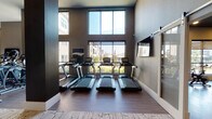 This single-story fitness center in Richmond, TX, is designed to support a comprehensive workout experience through several distinct functional zones. The space is characterized by soaring ceilings, neutral gray tones, and a combination of textured carpet and herringbone-patterned hardwood-style flooring that varies by area.

The cardio section is positioned along a wall of oversized windows that rise nearly to the ceiling, flooding the area with natural light and offering outdoor views of the surrounding greenery. Multiple treadmills, a recumbent bike, and additional cardio machines are arranged here, accompanied by wall-mounted televisions. A dedicated strength training area features cable machines, a Smith machine, free weights, kettlebells, and barbells with weight plates, all reflected in large framed mirrors that enhance the sense of space.

A separate studio room accommodates group fitness and cycling, equipped with spin bikes, stability balls, resistance bands, yoga mats, and a full-wall mirror with a mounted display. Additional amenities include a rack of medicine balls, plyo boxes, and a TRX suspension system. A built-in water fountain station is conveniently located within the main floor area, and glass-panel entry doors provide a sleek, modern transition into the space.