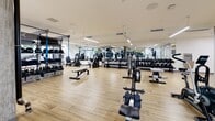 Located at 1530 Cassil Pl in Los Angeles, CA, this modern fitness facility is thoughtfully designed to support a wide range of workout styles and training needs. The main gym floor is expansive and bright, featuring light hardwood-style flooring, clean white walls, and rows of linear overhead lighting that create a well-lit, energetic environment. Exposed concrete columns add an industrial character throughout the space, complementing the contemporary aesthetic. The main floor is comprehensively equipped with treadmills, stationary and air bikes, rowing machines, cable systems, free weight racks with dumbbells, adjustable benches, and a functional training rig complete with kettlebells and suspension trainers. Full-length mirrors along the walls enhance the sense of openness and allow users to monitor their form during workouts. A dedicated studio room provides an open, flexible space for group classes or personal training sessions, outfitted with wall-mounted TVs, mirrors, stability balls, BOSU trainers, and plyometric boxes. An additional training area opens directly to a lush outdoor space, bringing in natural light and greenery, ideal for rehabilitation or functional movement work. The facility's seamless layout and high-quality equipment make it a well-rounded destination for fitness in the heart of Los Angeles.