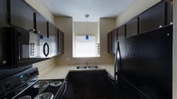 This single-floor kitchen is designed with a functional galley-style layout, making efficient use of the available space. Dark espresso-toned cabinetry lines both sides of the kitchen, providing ample storage from countertop height up to the ceiling. The lower cabinets are complemented by matching upper units, creating a cohesive, bold aesthetic against the neutral beige walls.

Light-toned countertops offer a contrast to the dark cabinetry, providing practical workspace on both sides of the kitchen. A double-basin stainless steel sink is positioned beneath a window fitted with white horizontal blinds, which allows natural light to brighten the space. A distinctive blue teardrop-shaped pendant light hangs from the ceiling above the sink area, adding a subtle decorative accent.

Black appliances, including a coil-burner electric range positioned in the foreground and a full-size refrigerator along the right wall, complete the kitchen's uniform color scheme. The ceiling features a textured popcorn finish. Overall, the kitchen presents a practical, well-organized space with a consistent dark-and-light design contrast.