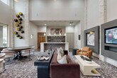 Located at 14698 Briar Forest Drive in Houston, TX, this single-floor game room is a sophisticated communal space designed for entertainment and relaxation. The room immediately impresses with its dramatic soaring ceilings, accented by stacked stone columns that rise from floor to ceiling, lending a refined, architectural character to the space. Exposed wooden ceiling beams and an eye-catching chandelier with amber globe pendants add warmth and visual interest overhead.

The lounge area is anchored by a plush velvet sofa and leather accent chairs arranged around a sleek coffee table, all set atop a richly patterned area rug. A dedicated media wall, finished in deep gray with recessed display niches housing curated artwork and a large flat-screen television, serves as a focal point for entertainment.

A stylish wet bar features mosaic tile cladding, a light stone countertop, pendant lighting, and a full-size wood-paneled refrigerator, making it well-equipped for hosting. A dining area with round tables and upholstered chairs provides additional gathering space. Large windows along the perimeter bring in abundant natural light and offer views of the surrounding greenery, completing the inviting, upscale ambiance of this well-appointed game room.