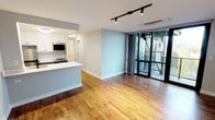This well-appointed single-floor unit offers a thoughtfully designed open-concept layout that seamlessly connects the kitchen and living areas. Rich hardwood flooring flows throughout the main living space, complemented by soft gray walls and crisp white trim that create a clean, contemporary aesthetic. Black track lighting runs along the ceiling, providing flexible, directional illumination across the open floor plan.

The kitchen is efficiently designed with white shaker-style upper cabinetry, contrasting gray lower cabinets, and a speckled quartz countertop that extends into a peninsula with an undermount sink. Stainless steel appliances — including a refrigerator, range, and over-the-range microwave — are paired with a classic white subway tile backsplash for a polished finish. A black matte faucet adds a modern accent.

The spacious living area opens directly onto a balcony through full-height black-framed glass sliding doors, allowing natural light to pour in and offering outdoor access. A ceiling-mounted cassette HVAC unit ensures year-round climate comfort.

The bathroom features a full soaking tub and shower combination, fully surrounded by large-format white tile in a stacked pattern. A wall-mounted vanity with a warm wood finish supports a white countertop and undermount sink, with an illuminated mirror completing the clean, modern look.