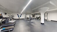 This fitness center is a comprehensively outfitted facility spread across a single floor, designed to support a wide range of workout routines. The space is anchored by a bold navy "W" logo mural on the white wall, lending the gym a distinct branded identity. Durable speckled rubber flooring runs throughout, providing both safety and resilience across all training zones.

The main strength area is generously sized, housing a variety of cable machines, weight benches, a free-weight rack stocked with dumbbells and kettlebells, and multi-functional cable stations. Structural columns with flared bases are integrated naturally into the layout without impeding usability. Linear LED strip lighting runs along the ceiling, ensuring bright, even illumination across the entire floor.

A separate cardio zone accommodates treadmills, stationary bikes, ellipticals, and a rowing machine, with a wall-mounted television for entertainment during workouts. An additional open functional training area features stability balls in multiple sizes mounted on the wall, a cable attachment station, and open floor space ideal for stretching or bodyweight exercises.

The facility also includes a water station, waste receptacles, and storage solutions for accessories. Small windows in select areas provide modest natural light, complementing the clean, white-walled interior and creating a practical, focused training environment.