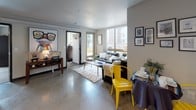 This well-appointed single-floor unit offers a thoughtfully designed layout that balances comfort and functionality. The open-concept living area is anchored by polished concrete floors that extend throughout, giving the space an urban, contemporary feel. Large windows flood the room with natural light and frame views of the surrounding neighborhood, while a sliding glass door leads to an outdoor balcony — perfect for enjoying fresh air. The living space comfortably accommodates a seating area and dining space, creating a versatile environment for both relaxing and entertaining.

The kitchen is efficiently laid out along a single wall, featuring dark espresso cabinetry, stone-look countertops, and a full suite of stainless steel appliances including a range, dishwasher, microwave, and refrigerator. Open upper shelving adds to the modern aesthetic while maximizing storage.

The bedroom is a bright, carpeted retreat with a full-height window offering treetop and neighborhood views, providing a peaceful and airy atmosphere. Two bathrooms serve the unit — one compact and sleek with a stone-finish countertop, and a second more spacious bathroom with dark cabinetry, a wide vanity mirror, and soft flooring. Together, the spaces form a cohesive, modern living environment with clean lines and practical design throughout.
