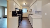 This single-floor residential unit offers a well-organized layout designed for practical, comfortable living. The galley-style kitchen is efficiently arranged with white cabinetry lining both sides, light countertops, black appliances including an electric range and refrigerator, a stainless steel sink, and wood-style flooring that extends through the main living areas. A window fitted with vertical blinds brings in natural light and adds a sense of airiness to the space.

The living area is generously proportioned, featuring dark wood-style flooring, neutral-toned walls, and a large multi-pane window that allows ample natural light and provides views of the surrounding exterior. Baseboard heating runs along the perimeter, ensuring year-round comfort. An entry door with hardware is conveniently positioned within the living space.

The bedroom is carpeted for warmth and comfort, and includes a louvered bi-fold closet door offering built-in storage. A window with vertical blinds provides natural light, and additional baseboard heating keeps the room comfortable. A second room with wood-style flooring and a double bi-fold closet offers flexible use as a bedroom or additional living space.

The full bathroom is clean and functional, featuring a white vanity, a combination tub and shower with tile surround, wood-style flooring, a frosted window, and brushed nickel fixtures throughout.