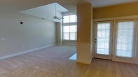 This single-floor residence offers a clean, open layout that maximizes both comfort and functionality. The entryway greets visitors with warm hardwood flooring and a glass-paneled door fitted with blinds, allowing natural light to filter in while maintaining privacy. A half-wall partition elegantly defines the transition between the entry and the main living area, adding architectural interest without sacrificing the sense of openness.

The living room is generously proportioned, featuring plush wall-to-wall carpeting that adds warmth underfoot. A ceiling fan is centrally mounted, providing both air circulation and a subtle design accent to the space. Large windows along the far wall flood the room with an abundance of natural light, creating a bright and airy atmosphere throughout the day. The neutral, warm-toned walls complement the light color palette, offering a versatile backdrop suited to a variety of interior styles.

The ceiling is notably elevated in the living area, contributing to a spacious, open feel. The crisp white trim and baseboards throughout provide a polished contrast to the soft wall tones, reinforcing the clean aesthetic of the home. Overall, this residence presents a well-lit, inviting environment with thoughtful architectural details that enhance everyday living.