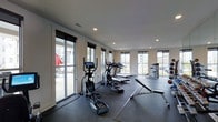 This fitness center offers a bright, open workout environment designed to support a variety of exercise routines. The space is finished with a dark rubberized flooring throughout, providing both durability and traction ideal for high-impact activities and weightlifting. Recessed lighting in the white ceiling complements the abundant natural light that streams in through multiple large windows and a sliding glass door along one wall, offering views of the surrounding outdoor area and creating an airy, energizing atmosphere.

The cardio zone is well-appointed, featuring a row of elliptical trainers and treadmills with integrated display screens, allowing users to track and customize their workouts. Adjacent to the cardio equipment, a flat workout bench and additional strength training equipment provide space for resistance and functional training exercises. The sliding glass door connects the interior to an outdoor area, enhancing ventilation and accessibility. The neutral wall tones and clean layout contribute to an organized, distraction-free environment suitable for focused fitness routines.