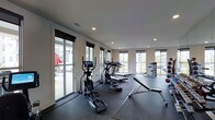 This fitness center offers a bright, open workout environment designed to support a variety of exercise routines. The space is finished with a dark rubberized flooring throughout, providing both durability and traction ideal for high-impact activities and weightlifting. Recessed lighting in the white ceiling complements the abundant natural light that streams in through multiple large windows and a sliding glass door along one wall, offering views of the surrounding outdoor area and creating an airy, energizing atmosphere.

The cardio zone is well-appointed, featuring a row of elliptical trainers and treadmills with integrated display screens, allowing users to track and customize their workouts. Adjacent to the cardio equipment, a flat workout bench and additional strength training equipment provide space for resistance and functional training exercises. The sliding glass door connects the interior to an outdoor area, enhancing ventilation and accessibility. The neutral wall tones and clean layout contribute to an organized, distraction-free environment suitable for focused fitness routines.