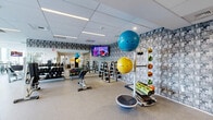 Located at 10 New Street in Boston's East Boston neighborhood, this fitness center offers a generously sized, single-floor exercise space designed to support a full range of workout routines. The room is bright and airy, thanks to expansive floor-to-ceiling windows that line the perimeter walls, flooding the interior with natural light and providing sweeping panoramic views of the Boston skyline and waterfront. The suspended tile ceiling with recessed linear lighting ensures consistent illumination throughout.

The exercise room is thoughtfully organized into distinct workout zones. A dedicated cardio area features a row of stationary bikes, treadmills, and elliptical machines positioned directly in front of the large windows, allowing users to enjoy the scenic views during their workouts. Wall-mounted flat-screen televisions add an entertainment option for cardio sessions. A strength training zone is equipped with a multi-station cable machine, adjustable benches, and a fully stocked dumbbell rack with a wide range of free weights. An additional functional fitness area includes stability balls, medicine balls, and a rowing machine.

A decorative textured feature wall adds a striking visual accent to the space. The durable, rubber-composite flooring throughout provides both comfort and safety. Glass partition walls and sliding doors connect the fitness center to an adjacent common area, enhancing the open, airy feel of the overall space.