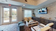 Located at 6805 Walnut Branch Lane in Charlotte, NC, this single-floor amenity space offers a thoughtfully designed environment for relaxation, entertainment, and social gathering. The layout encompasses several distinct zones, each with its own character and purpose.

The main lounge area is anchored by a textured stone accent wall adorned with wall-mounted flat-screen TVs, complemented by a built-in dark wood credenza with cabinetry below. Seating arrangements feature sleek, mid-century-inspired armchairs in neutral tones paired with low coffee tables and round upholstered ottomans. A patterned mosaic-style area rug adds visual depth, while sheer curtains filter natural light from large windows offering outdoor views.

A dedicated screening room — referred to as the "Reel Room" — provides theater-style seating with rows of cushioned armchairs arranged around small side tables, beneath a dramatic dark coffered ceiling with recessed lighting. Abstract artwork adorns the walls, enhancing the cinematic atmosphere.

Adjacent to the screening area, a game room features dining-height tables and chairs alongside a large board game display, connected via sliding barn doors. A starburst chandelier adds an elegant focal point overhead.

The space is unified by consistent flooring transitions between tile and patterned carpet, recessed and decorative lighting throughout, and a cohesive contemporary aesthetic that balances comfort with refined design.