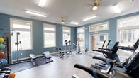 This single-floor fitness center offers a well-rounded workout environment designed to support a variety of exercise routines. The space is painted in a calming blue-gray tone, creating a focused yet inviting atmosphere for physical activity. Large white-trimmed windows line the walls, flooding the room with abundant natural light and providing pleasant views of the surrounding greenery outdoors.

The flooring consists of smooth, light-toned hardwood-style planks, with protective rubber mats placed beneath the free weight and strength training area. Equipment includes an adjustable weight bench with dumbbells, an elliptical cross-trainer, and multiple cardio machines such as treadmills and a stationary bike, catering to both cardiovascular and resistance training needs.

Overhead, ceiling fans with brass fixtures help maintain comfortable air circulation throughout the space. Recessed fluorescent lighting supplements the natural light, ensuring the room remains well-lit at all times. A mirrored wall panel near the entrance enhances the sense of space and allows users to monitor their form during workouts. A clearly marked exit door with brick surround accents provides easy access to the exterior. Overall, the fitness center is clean, functional, and thoughtfully arranged to maximize usability.