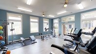 This single-floor fitness center offers a well-rounded workout environment designed to support a variety of exercise routines. The space is painted in a calming blue-gray tone, creating a focused yet inviting atmosphere for physical activity. Large white-trimmed windows line the walls, flooding the room with abundant natural light and providing pleasant views of the surrounding greenery outdoors.

The flooring consists of smooth, light-toned hardwood-style planks, with protective rubber mats placed beneath the free weight and strength training area. Equipment includes an adjustable weight bench with dumbbells, an elliptical cross-trainer, and multiple cardio machines such as treadmills and a stationary bike, catering to both cardiovascular and resistance training needs.

Overhead, ceiling fans with brass fixtures help maintain comfortable air circulation throughout the space. Recessed fluorescent lighting supplements the natural light, ensuring the room remains well-lit at all times. A mirrored wall panel near the entrance enhances the sense of space and allows users to monitor their form during workouts. A clearly marked exit door with brick surround accents provides easy access to the exterior. Overall, the fitness center is clean, functional, and thoughtfully arranged to maximize usability.