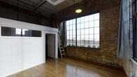 This single-floor space showcases a compelling industrial aesthetic rooted in its architectural heritage. The most commanding feature is the expansive grid-pane factory window set within a thick exposed brick wall, flooding the interior with generous natural light and offering open views of the surrounding cityscape. The original brick masonry spans the full height of the wall, lending warmth and textural character to the raw industrial atmosphere.

Hardwood flooring throughout adds a natural warmth that contrasts beautifully with the rugged brick and dark ceiling structure above. The ceiling retains its industrial character with visible structural elements and a skylight or ceiling panel that supplements the natural light from the large window.

A partial loft or elevated platform area is accessible via a freestanding wooden ladder, providing additional usable space or storage above the main floor. A private room or enclosure with a traditional paneled door and transom window sits adjacent to the loft structure, offering a more defined and enclosed area within the open-plan layout.

Overall, the space strikes a balance between raw industrial character and functional versatility, making it well-suited for creative, live-work, or commercial uses.