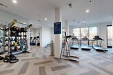 This well-appointed fitness center occupies a single, generously sized floor, offering a comprehensive workout environment designed with both functionality and aesthetics in mind. The space is organized into distinct zones, allowing users to move seamlessly between cardio, strength training, and free weight activities.

The main cardio area features a row of treadmills and elliptical machines positioned along a wall of large windows, providing natural light and outdoor views of the surrounding streetscape. Adjacent to this zone, a dedicated strength training section houses a selection of cable machines and selectorized equipment, supported by full-length mirrors that enhance the sense of space and assist users during exercise.

A separate free weights area is equipped with kettlebells, medicine balls, dumbbells, and benches, accompanied by additional mirrors and a punching bag for versatile training options. The flooring throughout the entire space features a distinctive herringbone-patterned carpet tile in neutral grey and beige tones, adding visual interest underfoot.

Crisp white walls are complemented by an accent wall finished in a deep teal-blue, introducing a modern color contrast. Recessed lighting throughout ensures even illumination, while the structural columns are cleanly integrated into the layout. Fitness center rules are prominently posted, and the space includes an accessible entry door leading to an outdoor area.