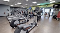 This fitness center offers a thoughtfully designed, single-floor layout built to accommodate a comprehensive workout experience. The cardio area is a standout feature, lined with rows of Life Fitness elliptical trainers and other cardio equipment, providing ample options for members to engage in low-impact or high-intensity aerobic exercise. Mounted televisions are positioned throughout the space, allowing members to stay entertained during their sessions. The flooring is a durable hardwood-style surface, combining practicality with a clean, polished aesthetic. The drop ceiling is outfitted with bright recessed panel lighting, ensuring the entire floor is well-illuminated. Accent walls in bold green and dark tones add visual energy to the environment, while motivational graphics and fitness-themed artwork contribute to an uplifting atmosphere. Large windows toward the rear of the space invite natural light and provide views to the outside, creating an open, airy feel. The overall layout is spacious and organized, with clear pathways between equipment for ease of movement. This facility is well-suited for members seeking a fully equipped, comfortable, and energizing space to pursue their fitness goals.