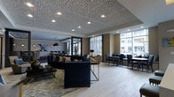 This elegantly designed single-floor space makes a strong first impression through its thoughtful blend of modern aesthetics and functional comfort. The most distinctive architectural feature is the ornate, patterned ceiling with an intricate geometric cutout design, complemented by recessed lighting that casts a warm, ambient glow throughout the space.

The open-plan layout is divided into distinct seating zones, each with its own character. A central lounge area anchors the space with deep navy blue sofas adorned with decorative throw pillows, arranged around a sleek gold-accented coffee table and a patterned area rug. Adjacent to this is a more intimate dining or casual seating area featuring dark chairs and small round tables, ideal for smaller gatherings or individual use.

Large floor-to-ceiling windows line the perimeter, flooding the interior with generous natural light and offering views of the surrounding urban environment. Sheer vertical drapes soften the light while maintaining an airy, open atmosphere. Light-toned hardwood flooring runs throughout, contrasting beautifully with the darker furnishings.

Pendant lighting fixtures hang near what appears to be a bar or service counter area in the background, adding a stylish focal point. The overall ambiance is upscale and welcoming, combining contemporary design elements with comfortable, functional furnishings suited for a communal or hospitality setting.