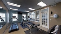 This fitness center offers a practical and well-organized space designed to accommodate a variety of workout needs. The room features a dark drop-panel ceiling with recessed fluorescent lighting that ensures consistent, bright illumination throughout. The walls are painted in a calming combination of teal and warm beige tones, contributing to an energizing yet focused atmosphere. Large windows along the perimeter flood the space with natural light and provide views into an adjacent lounge or common area through glass-paneled French doors, creating an open and connected feel. The flooring consists of durable, low-pile carpet with rubberized anti-fatigue mats positioned beneath the equipment for added safety and comfort. The fitness floor is equipped with multiple treadmills, an elliptical cross-trainer, and a weight bench, offering users a balanced selection of cardiovascular and strength training options. A wall-mounted television is positioned for convenient viewing during workouts. The layout is thoughtfully arranged to maximize the available floor space while maintaining clear pathways between equipment, ensuring ease of movement and a safe training environment suitable for everyday use.