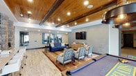 This single-floor clubhouse is thoughtfully designed to serve as a premier gathering and recreation space. Throughout, rich wood-paneled ceilings with exposed beam detailing create a warm, inviting atmosphere, complemented by recessed lighting that ensures the space is well-lit at all times.

The lounge area is furnished with a deep navy blue sofa, coordinating accent chairs, and a patterned area rug, all anchored by a wall-mounted flat-screen television. Colorful abstract artwork adorns the walls, adding a modern artistic touch. A billiards table positioned nearby makes this zone ideal for casual entertainment and social gatherings.

Adjacent to the lounge is a well-appointed kitchen and dining area that serves as the social hub of the clubhouse. A large central island with a light stone countertop and reclaimed wood detailing provides ample seating with bar-height stools. Dark cabinetry lines the walls alongside a second flat-screen television, while pendant lighting in brass and matte black finishes hangs above, adding industrial-chic character. Natural light streams in through large windows dressed with curtains, illuminating the stone accent columns and wood-look tile flooring. High-top dining tables complete the space, offering a relaxed café-style setting perfect for casual meals or social events.