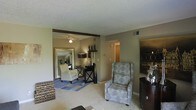 This single-floor space offers a warm and inviting layout designed for comfortable everyday living. The main living area is finished with neutral-toned carpeting and painted walls in a soft gray-beige palette, creating a calm and cohesive atmosphere throughout. A textured ceiling adds subtle character overhead, while a decorative exposed beam serves as an architectural accent that visually connects the spaces.

The living area is generously sized, accommodating a cushioned armchair upholstered in a blue-green pattern alongside additional seating, a dark wood dresser, and decorative wall art. A large floor mirror in an ornate gold frame enhances the sense of depth and light in the room.

An open passage leads into an adjoining sitting or study area, where a ceiling fan with a light fixture provides both air circulation and ambient lighting. This secondary space features a sleek writing desk with an X-base frame, a cream upholstered chair, and open shelving displaying decorative figurines, creating a thoughtful and functional nook.

Natural light filters through the space, complemented by warm artificial lighting. A doorway visible in the background hints at additional rooms beyond, suggesting a practical and well-connected floor plan. Overall, the space strikes a balance between elegance and functionality.
