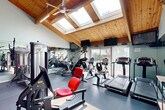 Located at 2010 W Jackson St in Morton, Illinois, this fitness center offers a thoughtfully designed exercise space that combines functional amenities with an inviting architectural character. The single-floor layout provides a comfortable and well-organized environment for a comprehensive workout experience.

The exercise room is defined by its striking vaulted ceiling, clad in warm natural wood planking and supported by exposed timber beams. A row of skylights runs along the roofline, flooding the space with abundant natural light throughout the day, while a ceiling fan ensures comfortable air circulation. Full-length mirrors line the walls, visually expanding the room and allowing users to monitor their form during exercise.

The facility is equipped with a wide selection of Nautilus and other brand-name strength machines featuring distinctive red upholstery, including cable machines, leg press, and seated resistance equipment. The cardio area includes multiple treadmills, elliptical trainers, and stationary bikes, all positioned to take advantage of the natural light and outdoor views through large windows. Mounted televisions provide entertainment during workouts.

A separate area with rubberized flooring and patterned carpet houses additional amenities, including exercise balls, free weights, and vending machines for post-workout refreshment. The durable rubber flooring throughout ensures safety and comfort underfoot, making this fitness center a well-rounded and welcoming space for daily exercise.