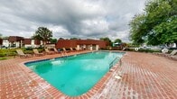Located at 6300 Old Canton Rd in Jackson, Mississippi, this outdoor amenity area centers around a generously sized rectangular swimming pool with clear, inviting turquoise water. The pool is accessed via dual stainless steel entry rails and is framed by a classic red brick deck that extends broadly on all sides, providing ample space for relaxation and movement. Surrounding the pool, numerous lounge chairs and sling-back seating offer comfortable spots for sunbathing or unwinding poolside. A tiled waterline detail and depth markers add a functional, well-maintained character to the pool itself. The entire area is enclosed by a black wrought iron fence, ensuring safety and privacy, with a posted lifeguard notice for added awareness. A poolside utility or amenity building with a distinctive brown metal roof sits adjacent to the pool, offering convenient access to facilities. Mature trees and well-tended landscaping, including flowering shrubs and greenery, border the space, providing natural shade and a pleasant visual backdrop. Umbrella-shaded seating stations are scattered throughout, adding to the casual, resort-like atmosphere. The surrounding community buildings, featuring matching brown rooflines, are visible beyond the fence, tying the outdoor amenity space cohesively into the broader property.