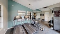 This fitness center is a comprehensively outfitted single-floor facility designed to support a full range of workout routines. The space is thoughtfully laid out to maximize both functionality and comfort, featuring a durable dark flooring surface suitable for high-traffic exercise use.

Cardio equipment is well-represented, with multiple True-brand treadmills and elliptical machines positioned near large, grid-paned windows that overlook an outdoor area, including what appears to be a pool. This placement allows natural light to fill the space while providing an engaging view during workouts.

For strength training, the facility includes a cable machine system, a multi-function weight apparatus, and several benches for targeted resistance exercises. The variety of equipment accommodates both upper and lower body training needs.

Full-length wall mirrors are installed throughout, enhancing the sense of openness and allowing users to monitor their form during exercise. The walls are painted in a calming combination of neutral beige and soft teal, contributing to an energizing yet comfortable ambiance.

Ceiling fans and recessed lighting provide consistent illumination and airflow throughout the space. A direct exterior door offers convenient access to the outdoor grounds. Wall-mounted amenity dispensers and posted instructional materials further enhance the usability of the facility.