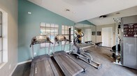 This fitness center is a comprehensively outfitted single-floor facility designed to support a full range of workout routines. The space is thoughtfully laid out to maximize both functionality and comfort, featuring a durable dark flooring surface suitable for high-traffic exercise use.

Cardio equipment is well-represented, with multiple True-brand treadmills and elliptical machines positioned near large, grid-paned windows that overlook an outdoor area, including what appears to be a pool. This placement allows natural light to fill the space while providing an engaging view during workouts.

For strength training, the facility includes a cable machine system, a multi-function weight apparatus, and several benches for targeted resistance exercises. The variety of equipment accommodates both upper and lower body training needs.

Full-length wall mirrors are installed throughout, enhancing the sense of openness and allowing users to monitor their form during exercise. The walls are painted in a calming combination of neutral beige and soft teal, contributing to an energizing yet comfortable ambiance.

Ceiling fans and recessed lighting provide consistent illumination and airflow throughout the space. A direct exterior door offers convenient access to the outdoor grounds. Wall-mounted amenity dispensers and posted instructional materials further enhance the usability of the facility.