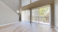 This two-story residence is designed with an open, airy feel, highlighted by dramatic double-height ceilings in the main living area. The living room features light-toned wood-style flooring and expansive floor-to-ceiling sliding glass doors fitted with vertical blinds, opening to a private balcony that overlooks a lush tree canopy, flooding the space with natural light.

The kitchen is appointed with gray shaker-style cabinetry, a white quartz peninsula countertop, a double undermount sink with a modern pull-down faucet, hexagonal tile backsplash, and stainless steel appliances, creating a clean, contemporary workspace.

A staircase with a decorative newel post leads to the upper level, where vaulted ceilings, carpeted flooring, and bright windows define the bedroom areas. The bedrooms are well-proportioned with neutral walls and good natural light. One bedroom features a walk-in closet with built-in shelving and hanging rods for ample storage.

The bathroom includes large-format tile flooring, gray vanity cabinetry with white countertops, and a framed mirror, presenting a cohesive, polished finish. Throughout, the neutral color palette and consistent finishes contribute to a bright, modern ambiance.
