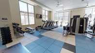 This modern fitness center is designed to provide a comprehensive workout experience in a bright, open environment. The space is anchored by a row of treadmills positioned to take full advantage of the large, floor-to-ceiling windows that line two walls, offering expansive views of the surrounding urban skyline and flooding the room with natural light. Ceiling fans help maintain a comfortable atmosphere during workouts.

The flooring features a distinctive multi-toned rubber tile layout in shades of blue, grey, and beige — a practical and durable surface well-suited for a high-activity environment. The free weight area is furnished with a dumbbell rack and an adjustable workout bench, ideal for a range of strength-training exercises. On the opposite side of the room, a cable and weight machine provides additional options for targeted muscle training, complemented by a stationary bike for low-impact cardio.

A wall-mounted flat-screen television is positioned for easy viewing from the cardio equipment, adding an entertainment element to the workout experience. The neutral-toned walls and clean, uncluttered layout contribute to a focused and functional ambiance, making this fitness center a well-rounded facility suited for both cardio and strength training routines.