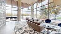 Located at 115 Christina Landing Dr in Wilmington, Delaware, this striking building lobby exemplifies contemporary architectural design. The space is defined by soaring floor-to-ceiling glass curtain walls that flood the interior with natural light and frame sweeping views of the Christina River waterfront and surrounding cityscape. Large-format stone tile flooring runs throughout, lending a polished, refined aesthetic underfoot.

The lobby is organized into distinct functional zones. A welcoming lounge area features a leather sofa and accent seating arranged atop an artfully patterned area rug, with elegant pendant lighting suspended from the double-height ceiling above. A secondary seating nook offers a pair of contemporary upholstered swivel chairs paired with a round marble side table, positioned beside a wood-paneled media wall with a wall-mounted display screen.

A dramatic backlit glass block feature wall serves as a bold artistic focal point near the entrance, with warm amber and cool tones creating a visually dynamic installation. The main entry vestibule opens through full-height glass doors, further reinforcing the transparency and openness of the design. A staffed reception counter with a stainless steel finish and glass shelving anchors the service area, leading into a corridor with access to additional spaces. Wood-paneled ceiling details and cylindrical columns complete the sophisticated, professional ambiance of this well-appointed lobby.