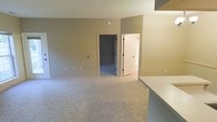 This single-story space presents a straightforward, well-maintained interior designed for practical living. The main living area is finished with wall-to-wall carpeting in a light neutral tone, providing a soft underfoot feel throughout. The walls are painted in a warm greige hue, creating a cohesive and calming atmosphere across the space. A textured ceiling adds a subtle design element overhead.

Two doorways are visible from the main living area, offering access to additional rooms. One doorway leads to a secondary carpeted room, likely a bedroom or additional living space, while the other opens to a brighter room that appears to include a closet, suggesting bedroom functionality with built-in storage. White door trim and frames provide clean contrast against the neutral walls.

A countertop edge visible in the foreground hints at an adjacent kitchen or bar area, indicating a practical transition between living and functional spaces. Electrical outlets are evenly distributed along the walls, and a smoke detector is centrally mounted on the ceiling. Overall, the space offers a clean, move-in ready environment with a simple, understated aesthetic suited to a variety of uses.
