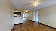 This single-floor space features an open-concept layout that seamlessly connects the living area and kitchen, creating a functional and cohesive environment. Rich hardwood flooring extends throughout the main living space, providing warmth and a sense of continuity across the floor plan.

The kitchen is positioned along one side of the open layout and is defined by dark espresso-toned cabinetry that contrasts with the light-colored countertops and walls. A white range and under-cabinet lighting add practicality to the cooking area, while a partial knee wall separates the kitchen from the living space without fully enclosing it.

Ceiling fans are installed in both the kitchen and living areas, contributing to air circulation and comfort throughout. The walls are painted in a neutral warm beige tone, complementing the flooring and cabinetry. Black baseboards provide a clean visual contrast along the perimeter of the rooms.

A doorway visible toward the rear of the space leads to an additional room, offering privacy and separation. Flush floor vents are present throughout, indicating a central HVAC system. The textured ceiling adds a classic finishing detail to the overall interior.