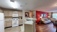 Located at 1070 Kenmore Dr in Clayton, NC, this single-story residence offers a well-designed, open-concept floor plan that maximizes both functionality and comfort. The kitchen is fully equipped with white appliances including a refrigerator, electric range, and built-in microwave, complemented by ample cabinetry in a neutral tone and a peninsula with a sink that opens directly to the living and dining areas. Wood-look flooring flows throughout the main living spaces, lending a cohesive and clean aesthetic. The living room features a bold red accent wall that serves as a striking focal point, with natural light entering through a large window. The adjacent dining area comfortably accommodates a full dining set and includes a wall-mounted wine rack for additional character. Two bedrooms are carpeted for comfort, each with ceiling fans, natural light from well-positioned windows, and generous closet space. The primary bathroom features wood-look flooring, a vanity with storage, and a shower-tub combination with built-in shelving. A second bathroom mirrors the same practical design. Textured ceilings are consistent throughout the home, and neutral wall tones in most rooms create a versatile backdrop suited for a variety of interior styles.