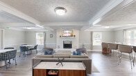 Located at 7266 Lockwood Ln in Indianapolis, IN, this single-floor communal space offers a thoughtfully designed open layout that seamlessly connects the living room, kitchen, and dining area. The interior is finished with warm hardwood-style flooring throughout, soft gray walls with crisp white wainscoting, and textured ceilings adorned with elegant coffered beamwork and crown molding that lend the space a refined character.

The expansive living room serves as the heart of the space, anchored by a classic white brick fireplace with a wall-mounted television above it. Multiple windows flood the room with natural light, while comfortable gray sofas and accent chairs with blue cushions create a welcoming, relaxed atmosphere. A marble-top coffee table and additional bar-height seating add to the versatility of the area.

The kitchen is equipped with rich wood cabinetry, dark countertops, a double sink, a range, dishwasher, and a full-size refrigerator, providing all the essentials for meal preparation. A convenient beverage cooler station is also accessible from the main living area.

The dining area features a round white table surrounded by patterned accent chairs, positioned near large windows and a glass-panel exterior door that invites in natural light and outdoor views, completing this functional and inviting communal space.
