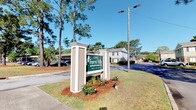 Forest Hills Apartments is a residential community located at 12012 Middleground Rd in Savannah, Georgia, 31419. The community is set against a backdrop of tall, mature pine trees that lend a natural, serene atmosphere to the property. The entrance is marked by a well-maintained monument sign framed by white columns and decorative landscaping, immediately establishing a sense of pride and curb appeal. The grounds are neatly kept, featuring manicured lawns, mulched planting beds with colorful flowers, and open paved roadways that wind through the community. The residential buildings are single-story structures with neutral-toned exteriors that blend harmoniously with the surrounding natural environment. Ample surface parking is available throughout the community, providing convenient access for residents. The tree-lined setting creates a peaceful, shaded environment, offering a sense of privacy and seclusion while remaining accessible to the broader Savannah area. The overall ambiance of Forest Hills Apartments reflects a quiet, well-established community that combines comfortable residential living with the natural beauty characteristic of coastal Georgia.