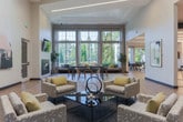 Located at 1800 Deerfield Pt in Milton, GA, this single-floor community clubhouse delivers a bright, contemporary atmosphere designed for both relaxation and productivity. The space is anchored by a dramatic lounge area with soaring floor-to-ceiling windows that flood the interior with natural light and frame lush, tree-lined outdoor views. Light hardwood flooring runs throughout, lending warmth and visual continuity across the open layout.

The main social area features a pool table, a modern fireplace with a mounted display above it, and curated artwork on the walls, creating an inviting yet stylish environment. A dedicated lounge nook with a vibrant yellow sofa and accent chairs sits beneath expansive gridded windows, offering a comfortable spot to unwind. Adjacent to this, a co-working zone provides seating and workspace for residents.

The kitchenette area is well-appointed with a large white waterfall-edge island, bar-height seating, warm wood-toned backsplash, and a beverage cooler—ideal for casual gatherings. A glass-door passage leads to a fitness room, visible from the main hall, keeping wellness accessible and integrated into the overall design. The space also connects to an outdoor patio area, further extending its functionality for community events and everyday use.