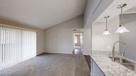Located at 601 W Renner Rd in Richardson, TX, this two-bedroom, one-bathroom residence offers a well-organized single-story layout with a cohesive, neutral color palette throughout. The combined kitchen and living area serves as the heart of the home, where striking vaulted ceilings lend an open, airy feel to the living space, which features plush carpeting and a view down the hallway connecting the rest of the unit. The kitchen is efficiently designed with dark wood-tone flooring, white cabinetry, granite countertops, and stainless steel appliances including a refrigerator and gas range. Two hallways provide smooth connectivity between living and private areas. The primary bedroom is generously sized and includes a walk-in closet, while the second bedroom also features its own closet and a window with natural light. Both bedrooms are finished with soft carpet. The bathroom is well-appointed with a granite-topped vanity, white cabinetry, a full soaking tub with shower, and wood-tone flooring that ties cohesively to the kitchen. Ceiling fans are present in the bedrooms for added comfort. The overall design emphasizes practicality and clean, modern finishes in a compact and efficient floor plan.