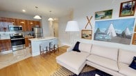 This single-floor space showcases a seamless open-concept layout that effortlessly connects the kitchen, dining, and living areas. The kitchen is outfitted with stainless steel appliances, warm wood cabinetry, and a sleek white countertop island with bar seating — ideal for casual meals or entertaining. Pendant lighting above the island adds a refined yet approachable touch to the space.

The living area is anchored by a generous light-toned sectional sofa, complemented by a bold floor lamp that provides both function and style. Light hardwood floors run throughout, lending warmth and continuity to the open floor plan. A patterned area rug defines the seating zone, adding texture and visual interest.

The walls are adorned with a curated selection of vintage-inspired artwork, including landscape paintings and decorative crossed oars, evoking a relaxed, outdoorsy aesthetic. The overall color palette — soft whites, warm woods, and muted tones — creates a bright, airy ambiance throughout.

A hallway visible from the main living area suggests additional rooms beyond, providing practical separation between the common areas and private spaces. The overall design balances modern convenience with a cozy, character-filled atmosphere.