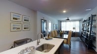 This single-floor residence showcases a thoughtfully designed open-concept layout that seamlessly connects the kitchen, dining, and living areas into one cohesive, flowing space. The kitchen is outfitted with a large center island topped with crisp white countertops, a deep stainless steel undermount sink with a sleek arc faucet, and dark cabinetry that provides a striking contrast. The island serves as both a functional workspace and a natural gathering point. Adjacent to the kitchen, the dining area accommodates a full-sized table with upholstered chairs, ideal for everyday meals or entertaining. A large decorative mirror adds depth and dimension to the space. The living room beyond is generously sized and filled with natural light streaming through expansive windows dressed with sheer curtains. A ceiling fan anchors the room, enhancing both comfort and airflow. Rich dark hardwood flooring runs throughout, grounding the space with warmth and elegance. The walls are finished in a clean, neutral tone that complements the overall modern aesthetic. Framed coastal artwork adorns the walls, adding a refined decorative touch. The open sightlines and abundant natural light create an inviting, airy ambiance throughout.