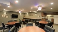 This single-floor communal space is thoughtfully designed to accommodate a variety of social and functional needs. The room features a combination of round dining tables with cushioned black chairs and a raised counter-height bar along the back wall, offering diverse seating options for both casual conversation and focused work. Warm wood tones from the tabletops and cabinetry contrast elegantly with the muted sage-green walls, creating a relaxed yet polished atmosphere. Overhead, decorative ceiling fans with ambient accent lighting complement a series of recessed lights, ensuring the space is well-lit throughout. Wall-mounted sconces add additional warmth and depth to the room. A large flat-screen television is mounted on one wall, making the space suitable for presentations or entertainment. Subtle wall art and decorative accents contribute to the inviting character of the room. A small kitchenette area is visible along one side, providing convenient access to refreshments. The neutral-toned carpet ties the design elements together, resulting in a cohesive and comfortable environment ideal for lounging, dining, or community gatherings.
