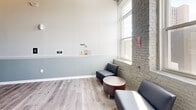 Located at 810 Monocacy St in Bethlehem, PA, this hallway space masterfully combines industrial heritage with contemporary design. The corridor is defined by whitewashed brick and concrete block walls that lend a raw, textural character throughout, while wood-look luxury vinyl plank flooring adds warmth and a polished finish underfoot. Classic white wainscoting with a soft gray paint treatment above creates a refined contrast against the rugged masonry walls.

Natural light filters in through tall, generously sized windows, illuminating the space and enhancing its open, airy feel. The soaring ceiling height adds a sense of grandeur rarely found in a transitional corridor. A stainless steel elevator serves as a sleek, functional focal point, framed by a bold dark header beam that reinforces the industrial aesthetic.

At one end, a small seating area furnished with modern charcoal chairs and a round wood-top table offers a comfortable waiting or gathering spot. A navy blue exit door with a glass panel provides clear wayfinding, complemented by accessible electrical panels and safety signage. The overall ambiance is clean, purposeful, and thoughtfully designed — balancing the building's historic character with the demands of a modern, accessible space.